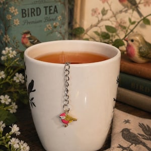 May include: A white ceramic mug filled with tea, featuring a bird-shaped tea infuser. The infuser is a small, colorful bird charm hanging from a chain. A tin of "BIRD TEA Premium Blend" is in the background, along with a bird-themed print and a decorative bird figurine.