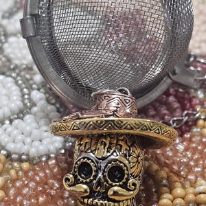 May include: A silver tea infuser with a sugar skull design. The skull is wearing a gold and copper sombrero. The infuser is sitting on a bed of colorful beads.