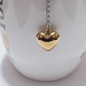 Könnte beinhalten: Ein goldener herzförmiger Tee-Infuser mit einer silbernen Kette, die am Rand einer weißen Teetasse hängt.