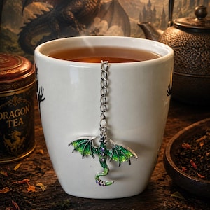 以下が含まれることがあります： 紅茶が入った白い陶器製のマグカップ。半透明の翼と小さな紫色のアクセントが特徴の緑色のドラゴンの形をした銀色の金属製ティーインフューザーが付いています。ドラゴンはマグカップの縁に掛かる銀色のチェーンに取り付けられています。