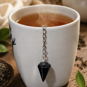 May include: A white ceramic mug filled with steaming hot tea. A silver-toned metal chain hangs over the rim of the mug, attached to a smooth, black, faceted, cone-shaped stone pendant that rests against the exterior wall of the white cup.