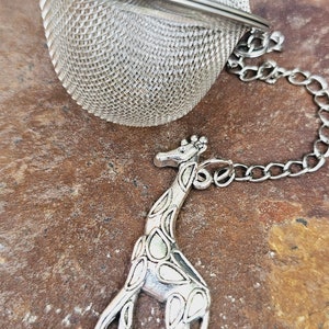 May include: A silver tea infuser shaped like a ball with a mesh design and a chain attached to a giraffe-shaped charm.
