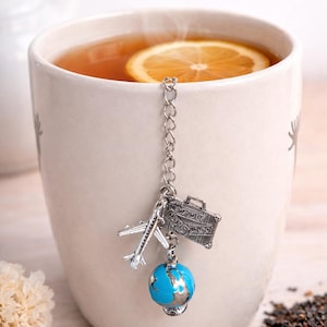 May include: A white mug filled with tea and a slice of orange. A travel-themed tea infuser with a silver airplane, suitcase, and globe hangs inside the mug. The suitcase has the words "NEW YORK PARIS TOKYO" on it.