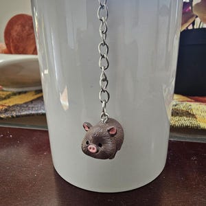 May include: A white ceramic mug with a decorative pig tea infuser. The pig is brown with pink accents and hangs from a silver chain. The mug is on a dark brown surface.
