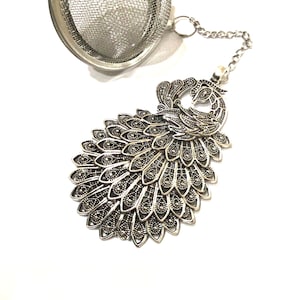May include: Silver tea infuser with a peacock design. The infuser has a round mesh ball with a chain attached to a peacock-shaped handle.