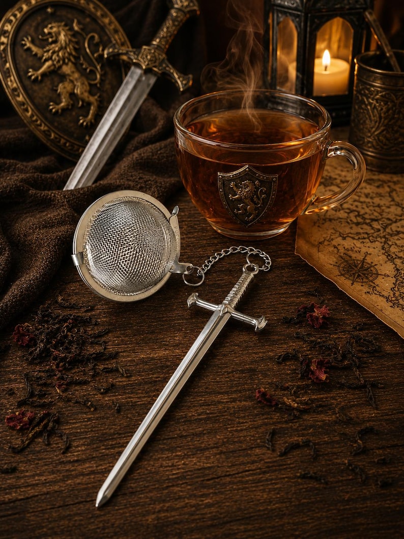 May include: A silver-colored metal tea infuser featuring a spherical mesh basket attached to a miniature sword-shaped handle by a small chain. The infuser is placed next to a glass teacup with a lion crest emblem and a sword-themed decor.