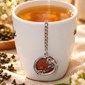 May include: A white ceramic mug filled with amber tea, featuring a silver-toned metal tea infuser chain. The infuser charm is a circular, glittery brown sandstone cabochon surrounded by a detailed silver-colored dragon figure.