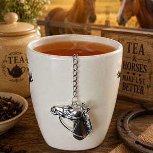 Peut inclure: Une tasse en céramique blanche remplie de thé fumant, dotée d'un infuseur à thé en métal argenté en forme de tête de cheval attaché à une chaîne. La tasse est décorée de texte et de graphismes noirs, dont la phrase TEA & HORSES MAKE LIFE BETTER.