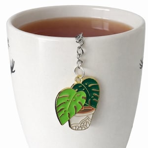 May include: A white ceramic mug filled with tea, featuring a decorative plant-shaped tea infuser. The infuser is a green and white enamel design of a potted plant with a gold-colored trim, suspended by a silver chain.
