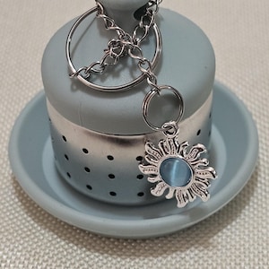 May include: A light blue ceramic tea infuser with a silver chain and a silver sun charm with a blue glass center. The infuser has a strainer on the bottom and sits on a matching saucer.