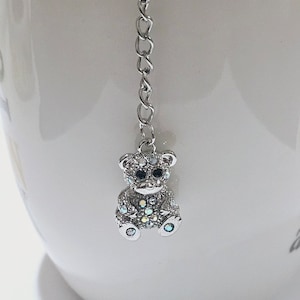 Puede incluir: Un infusor de té de plata con un dije de oso incrustado de cristales. El oso tiene ojos negros y está sentado en una taza de cerámica blanca.