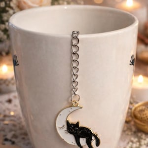 May include: A white ceramic mug with a decorative tea infuser. The infuser features a crescent moon design with a sleeping black cat. The infuser is attached to a silver chain. The mug has a floral design on the side.