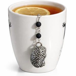 May include: A white ceramic mug with a slice of lemon in tea. A silver-colored tea infuser shaped like a brain hangs from the rim. The infuser has black lava beads on the chain.
