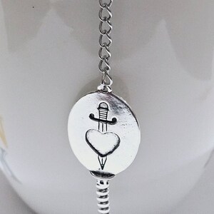 Op de afbeelding: Zilveren thee-infuser met een ketting en een ronde bedel met een hart dat door een dolk wordt doorboord.