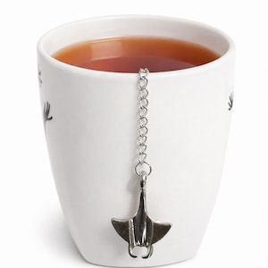 以下が含まれることがあります： お茶が入った白いセラミックマグカップ。銀色のオニイトマキエイのティーインフューザーがチェーンで吊り下げられています。マグカップは黒の装飾要素を備えたミニマリストデザインです。お茶は琥珀色です。