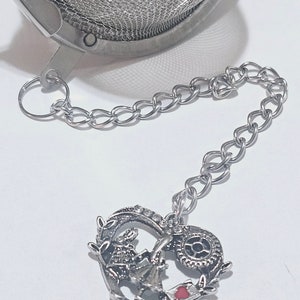 May include: A stainless steel tea infuser with a chain and a decorative heart charm. The charm features a rabbit, a clock, and a red heart. The infuser is designed for steeping loose leaf tea.