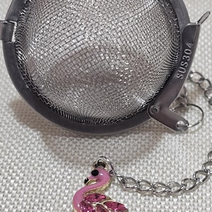 Puede incluir: Un infusor de té plateado con una bola de malla fina y una cadena unida a un amuleto de flamenco rosa con pedrería. El infusor está etiquetado como "SUS304".
