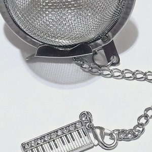 May include: Silver tea infuser with a chain and a piano-shaped charm. The infuser is a sphere with a fine mesh and a hinged lid. The charm has a white piano keyboard with black keys and clear rhinestones.
