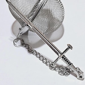 May include: A silver-colored metal tea infuser featuring a spherical mesh basket and a decorative handle shaped like a longsword. A small metal chain connects the sword-shaped handle to the mesh basket, which is designed for steeping loose tea leaves.
