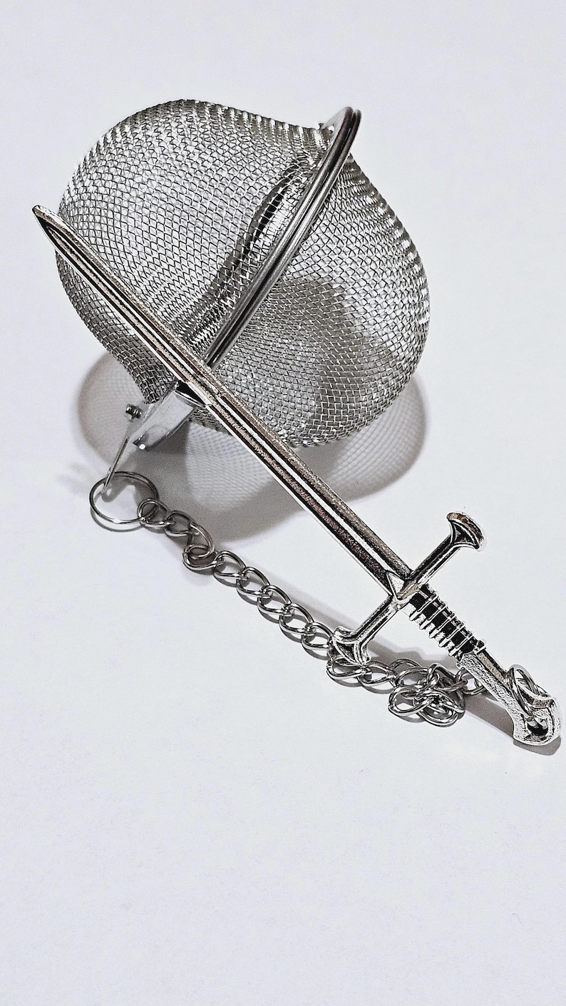 May include: A silver-colored metal tea infuser featuring a spherical mesh basket and a decorative handle shaped like a longsword. A small metal chain connects the sword-shaped handle to the mesh basket, which is designed for steeping loose tea leaves.