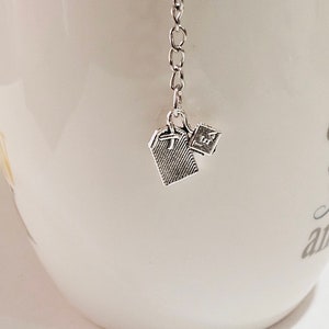 May include: A silver tea infuser with a chain and two tea bag charms. The charms are shaped like tea bags and have a textured surface.