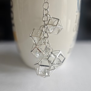 Puede incluir: Una cadena de plata con varios abalorios en forma de cubo llenos de cristales que cuelgan de ella. La cadena cuelga del borde de una taza de cerámica blanca.