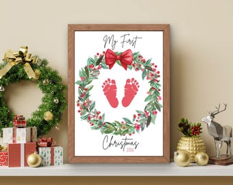 May include: A framed print featuring a watercolor Christmas wreath with green leaves and red berries, a red bow at the top, and two red baby footprints in the center. The text reads My First Christmas 2026 in a black script font on a white background.