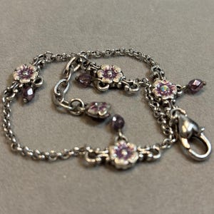 May include: Silver-toned bracelet featuring small, detailed purple flower charms and dangling beads. The bracelet has a delicate chain design and a secure clasp. A charming piece of jewelry.