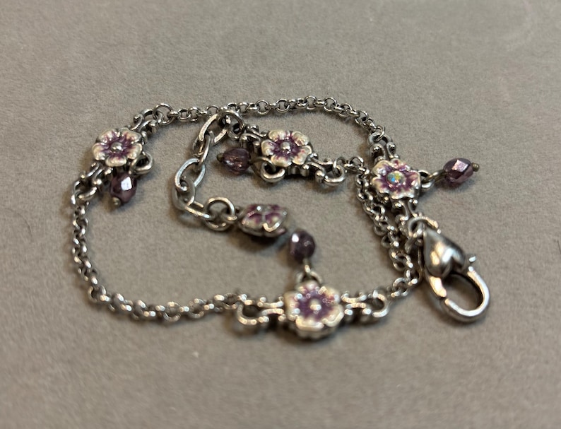 May include: Silver-toned bracelet featuring small, detailed purple flower charms and dangling beads. The bracelet has a delicate chain design and a secure clasp. A charming piece of jewelry.