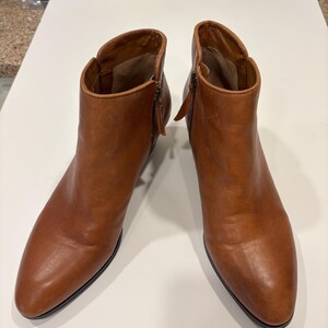 May include: A pair of brown leather ankle boots with side zippers. The boots have a pointed toe and a low heel. The leather has a slightly distressed appearance. The boots are displayed on a white surface.