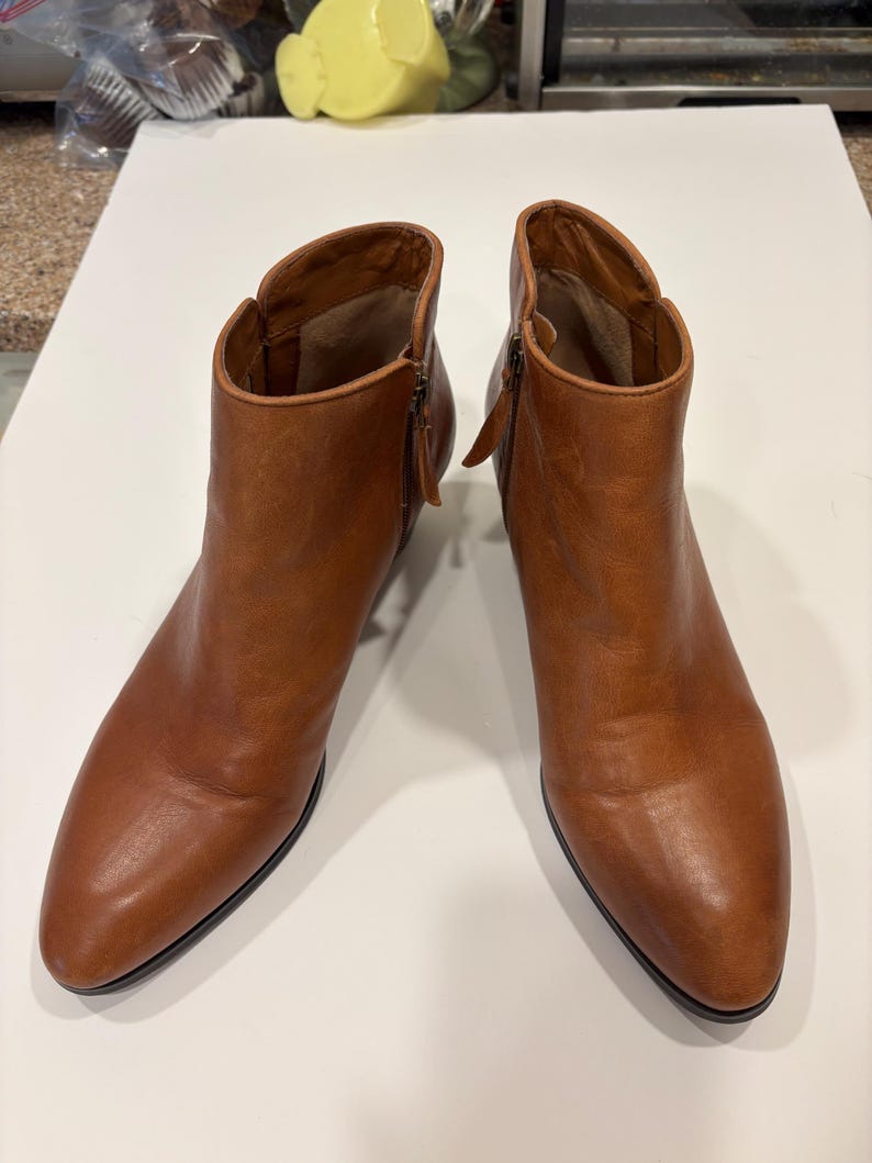 May include: A pair of brown leather ankle boots with side zippers. The boots have a pointed toe and a low heel. The leather has a slightly distressed appearance. The boots are displayed on a white surface.
