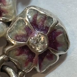 May include: Close-up of a silver-toned floral brooch. The flower has purple and green enamel petals and a clear, round gemstone center. The brooch is detailed and appears to be vintage or antique.