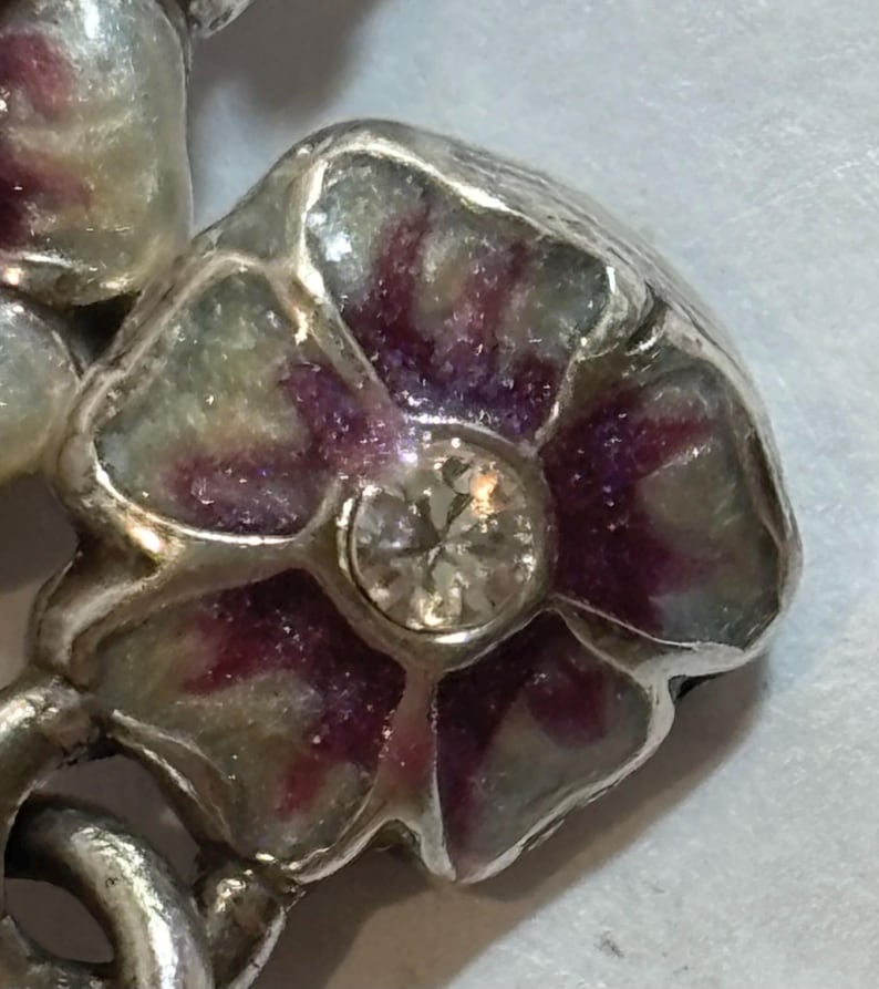 May include: Close-up of a silver-toned floral brooch. The flower has purple and green enamel petals and a clear, round gemstone center. The brooch is detailed and appears to be vintage or antique.
