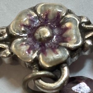May include: Close-up of a silver-toned floral charm with a purple and white enamel flower. The charm is connected to a silver ring and a faceted purple bead. The design is detailed and ornate.