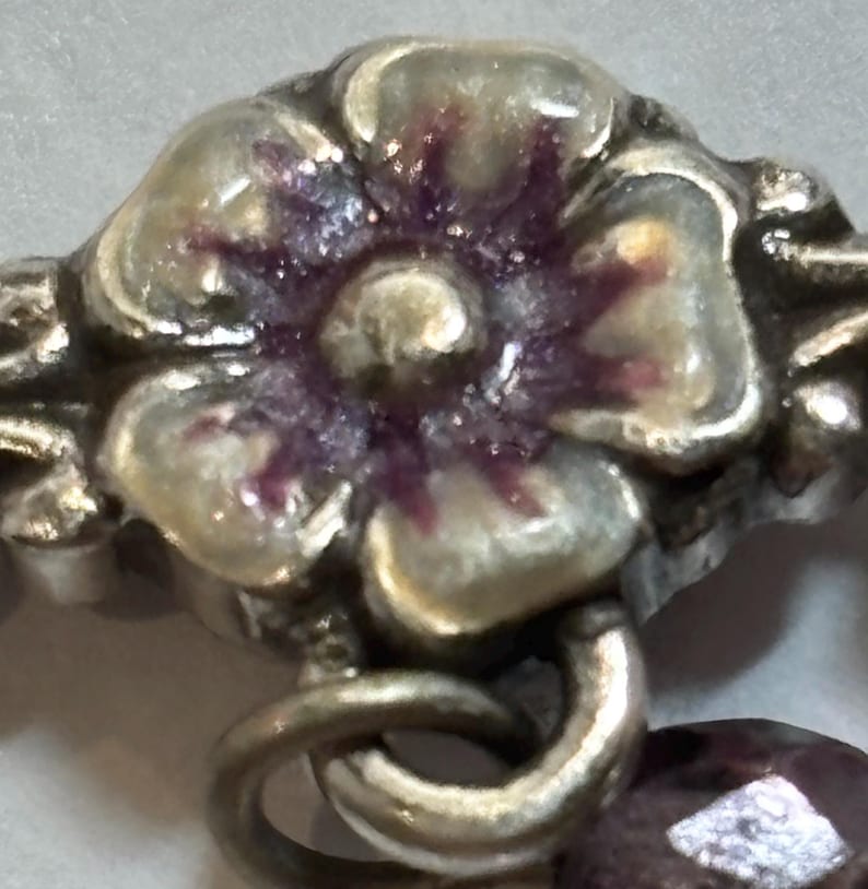 May include: Close-up of a silver-toned floral charm with a purple and white enamel flower. The charm is connected to a silver ring and a faceted purple bead. The design is detailed and ornate.