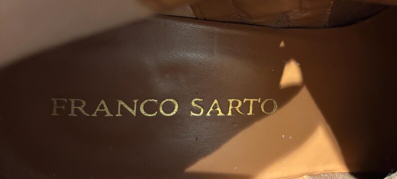 May include: Close-up of the interior of a brown leather shoe. The brand name "FRANCO SARTO" is embossed in gold lettering on the insole. The leather has a smooth, worn appearance.