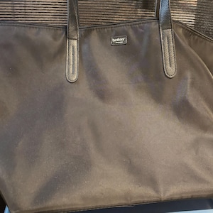 May include: A black tote bag with leather handles and trim. The bag has a rectangular shape and a large main compartment. The brand name "botkier" is visible on a small tag near the top. The bag is made of a smooth, possibly nylon, material.