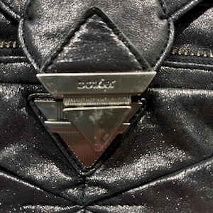 May include: A black quilted handbag featuring a prominent triangular silver-toned metal clasp closure. The metal hardware is engraved with the text "boker" across the top section of the triangular latch mechanism, set against the textured black material.