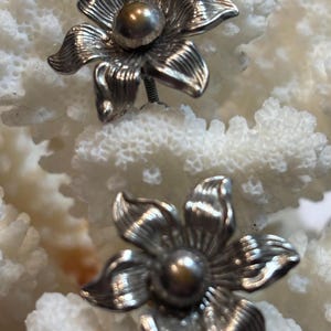 以下が含まれることがあります： 中央に真珠のようなビーズが付いた、2つの銀色の花のスタッドピアス。ピアスは、滑らかな金属と多孔質の表面のコントラストを生み出す、質感のある白い背景に対して配置されています。