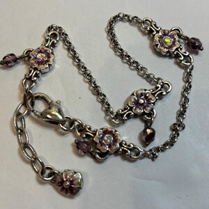 May include: Silver-toned bracelet featuring floral accents with pink enamel and iridescent centers. The bracelet includes a chain with a heart-shaped clasp and dangling purple beads. A delicate piece of jewelry.