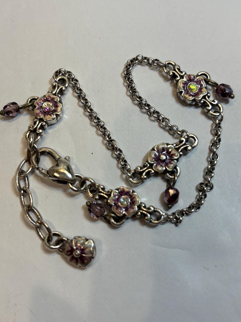 May include: Silver-toned bracelet featuring floral accents with pink enamel and iridescent centers. The bracelet includes a chain with a heart-shaped clasp and dangling purple beads. A delicate piece of jewelry.