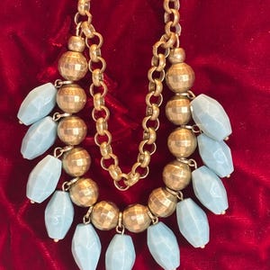 May include: A gold chain necklace with a row of large, faceted, light blue beads hanging from it. The necklace is on a red velvet background.