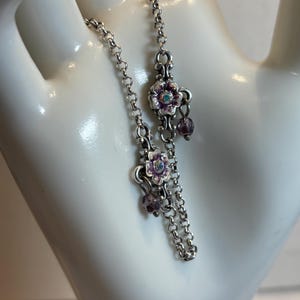 May include: Silver-toned bracelet with a delicate chain design. The bracelet features floral charms with purple and white enamel, accented with small purple beads and a central iridescent stone. A charming piece of jewelry.