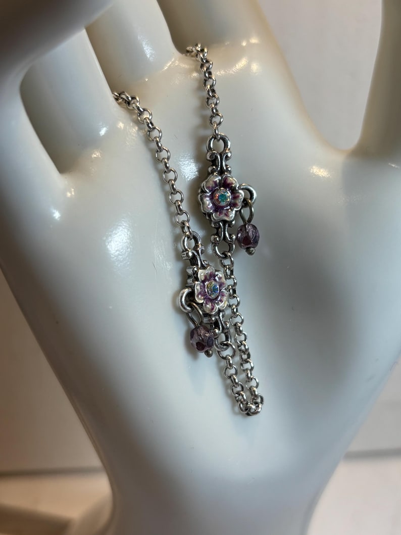 May include: Silver-toned bracelet with a delicate chain design. The bracelet features floral charms with purple and white enamel, accented with small purple beads and a central iridescent stone. A charming piece of jewelry.