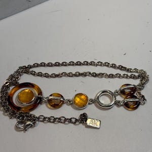 May include: Silver-toned necklace featuring a chain and decorative elements. The necklace includes amber-colored, circular, and round accents. The necklace has a lobster clasp and a small tag with the letters "AK".