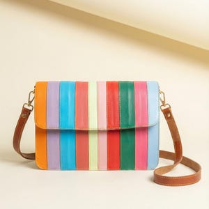 May include: A multi-coloured crossbody bag with a rainbow striped design. The bag features vertical stripes in various colours, including orange, purple, blue, pink, red, and green. It has a brown leather strap and gold-coloured hardware.