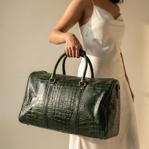 May include: A dark green leather duffel bag with a crocodile skin texture. The bag has two top handles and a zip closure. The bag is held by a person wearing a white dress.