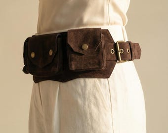 May include: A brown suede fanny pack with multiple pockets and a brass buckle. The belt bag is worn around the waist and has a rectangular shape with rounded edges. The pockets have brass button closures. The belt is worn over a cream-colored top and pants.