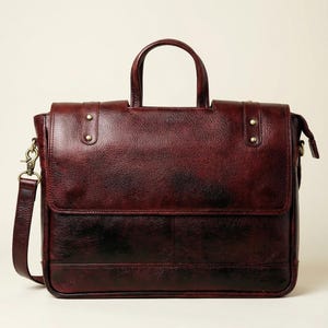 May include: A burgundy leather briefcase with a top handle and a detachable shoulder strap. The bag features a flap closure, brass-colored rivets, and a two-tone color gradient from burgundy to black.