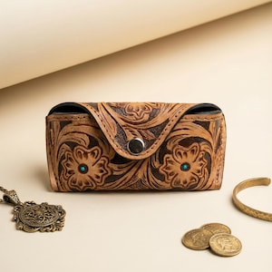 May include: A brown leather glasses case with floral tooling and a snap closure. The case has turquoise accents and is next to a gold bracelet, a pendant necklace, and coins.